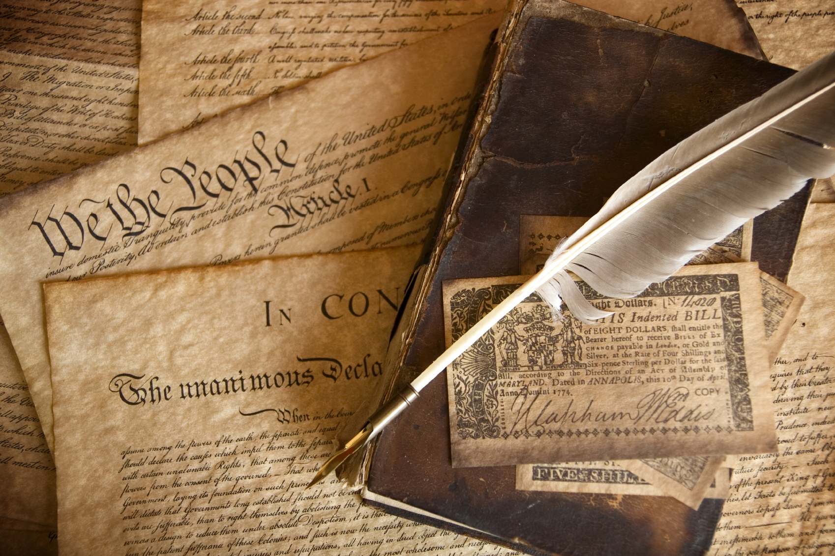 A photo of a pile of historical documents, including the Constitution, and the Declaration of Independence, as well as an aged book and a quill feather pen.