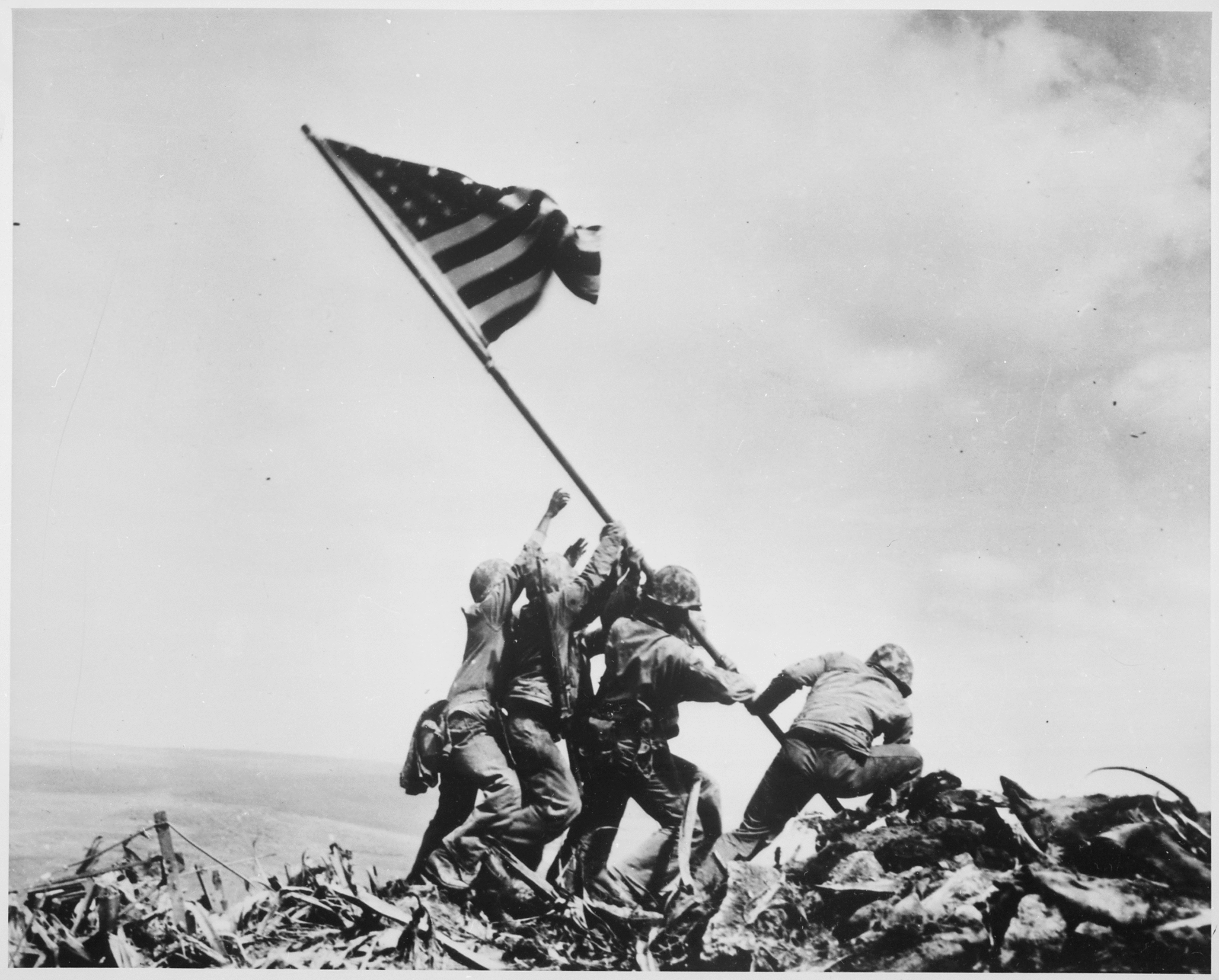 Four soldiers raise a US flag on a rocky hill of Iwo Jima overlooking the ocean.